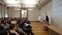 Abgebildet ist ein größerer Raum, links sieht man vier Sitzreihen voller Menschen, rechts der Referent Paul Downes aus Irland, der beim Pult steht und mittels Laptop seine Powerpoint-Präsentation präsentiert, im Hintergrund zu sehen ein Flipchart-Ständer