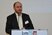 Abgebildet ist der Referent Dr. Nestor Kapusta (Medizinische Universität Wien), mit schwarzem Sakko und weißem Hemd, der sprechend hinter dem Rednerpult steht. Er trägt außerdem einen rötlichen Bart und eine schmale Brille.